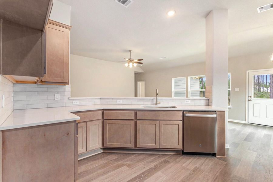 This photo showcases a modern kitchen with light wood cabinets, sleek countertops, and stainless steel appliances. It features a spacious open layout with a view into the living area, complemented by hardwood flooring and abundant natural light from large windows and a glass door.