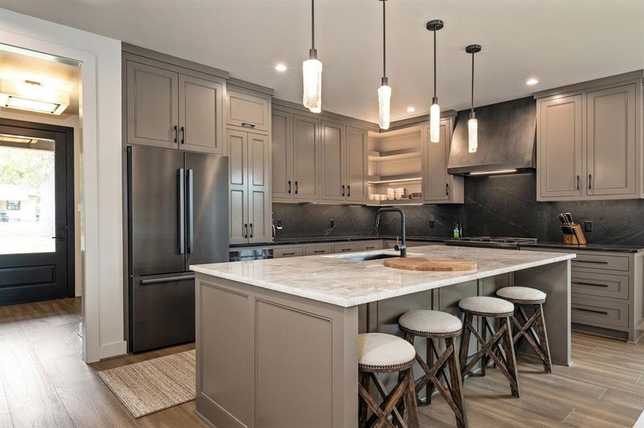 Kitchen with hardwood / wood-style floors, custom exhaust hood, stainless steel appliances, light stone counters, and an island with sink