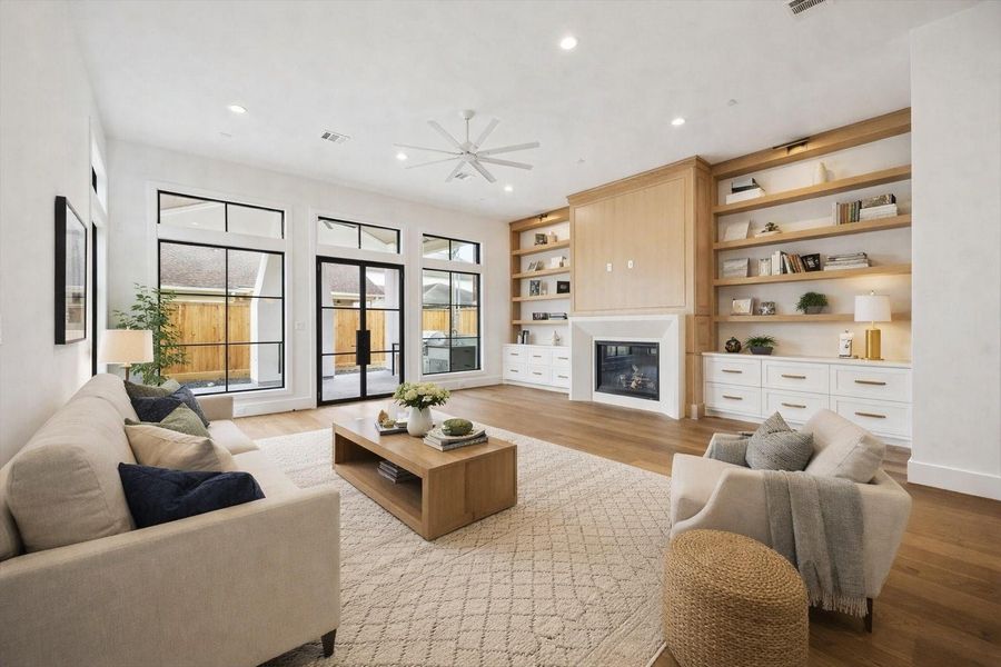 Open concept living room highlighted by floor to ceiling custom built ins, a sleek gas fireplace, and a mounted media wall. Expansive black framed windows and French doors invite natural light and seamless indoor outdoor flow, while wide plank hardwood floors and recessed lighting elevate the modern architectural design.  *Virtually Staged*