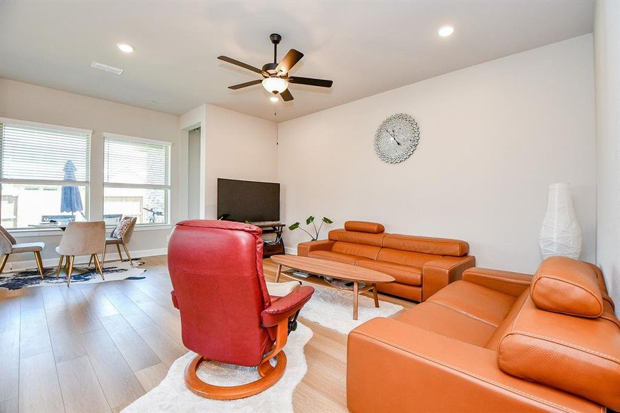 Bright and modern living room with light wood flooring, featuring a ceiling fan, stylish orange sofas, and a large wall clock. The space includes a cozy dining area by large windows, offering plenty of natural light.