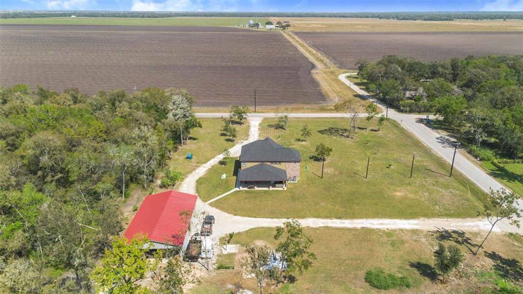 This aerial view shows a rural property with a house and a red-roofed outbuilding on a spacious lot. It is surrounded by open fields and bordered by a few trees, offering a peaceful countryside setting with plenty of open space.