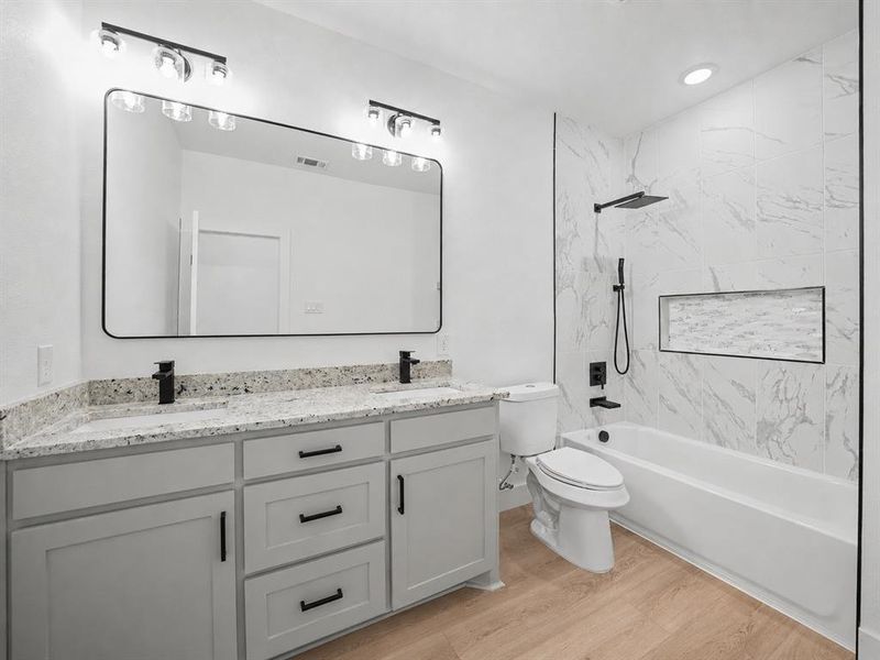 Bathroom featuring a double vanity with granite-style countertops, light wood-finish flooring, a full bathtub with a shower niche, and matte black fixtures Bathroom featuring a double vanity with granite-style countertops, light wood-finish flooring, a full bathtub with a shower niche, and matte black fixtures