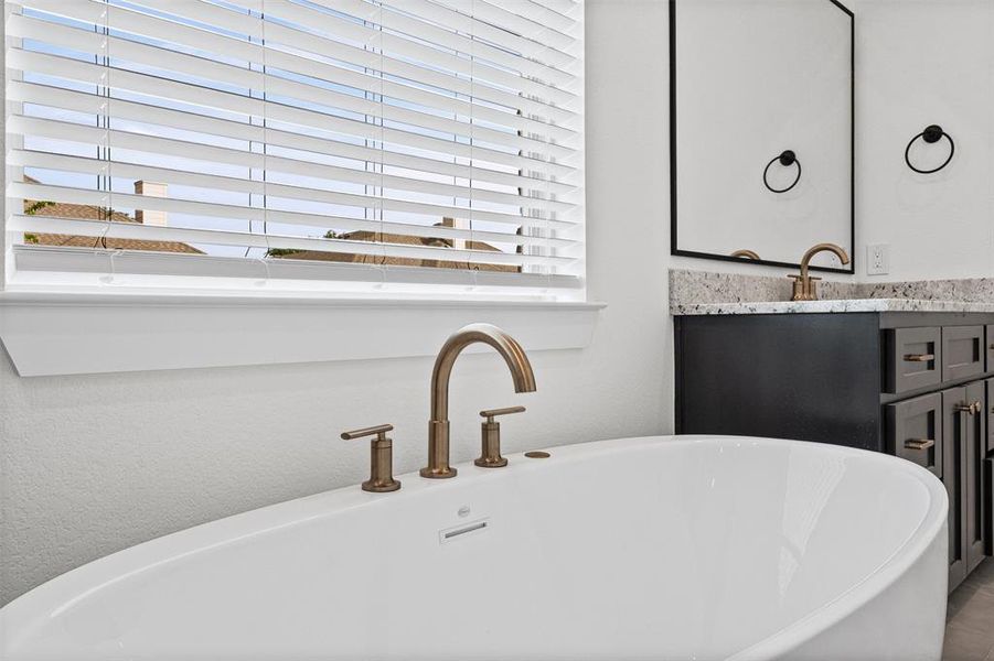 The bathroom includes a white freestanding bathtub with a bronze faucet, a double vanity with a dark wood cabinet, a granite countertop, and bronze fixtures, and a window with white blinds