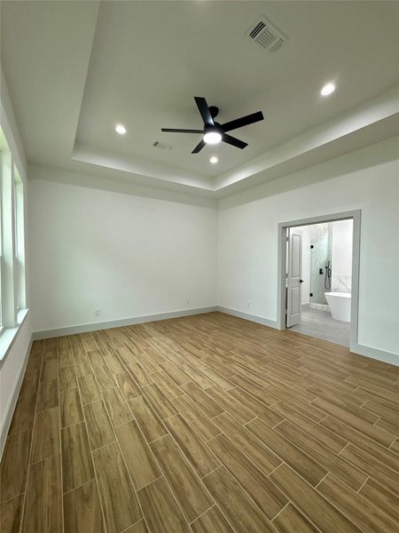 Spacious bedroom with wood-look tile flooring, tray ceiling with recessed lighting, and a modern ceiling fan. It features large windows for natural light and an open doorway leading to a luxurious bathroom with a freestanding tub. Spacious bedroom with wood-look tile flooring, tray ceiling with recessed lighting, and a modern ceiling fan. It features large windows for natural light and an open doorway leading to a luxurious bathroom with a freestanding tub.
