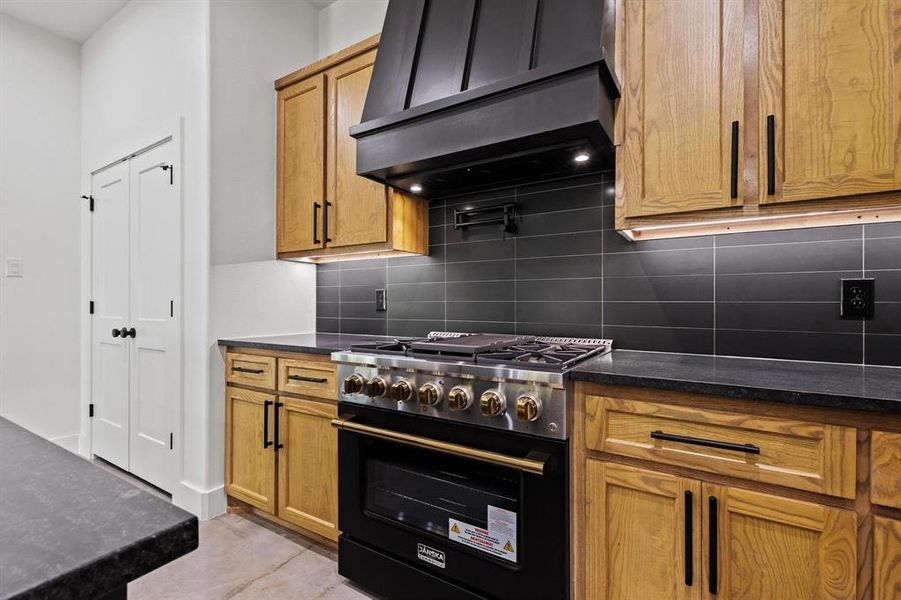 Kitchen with high end black range, range hood, backsplash, brown cabinets, and dark stone countertops