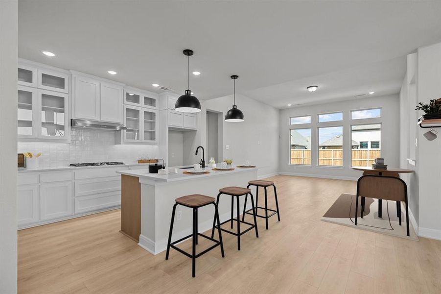 Modern kitchen featuring white cabinetry, a central island with countertop seating, stainless steel appliances, and wood-finish flooring extending into the open concept space with multiple windows Modern kitchen featuring white cabinetry, a central island with countertop seating, stainless steel appliances, and wood-finish flooring extending into the open concept space with multiple windows