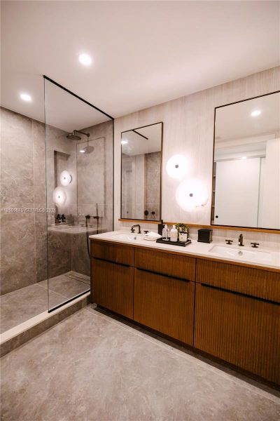 Custom double-sink vanity with fluted wood-finish cabinetry, frameless glass walk-in shower with floor-to-ceiling stone-look tiling, matte black fixtures, and dual wall-mounted vanity mirrors with integrated lighting. Images are for finish purposes only. Please refer to the floor plan for the layout. Custom double-sink vanity with fluted wood-finish cabinetry, frameless glass walk-in shower with floor-to-ceiling stone-look tiling, matte black fixtures, and dual wall-mounted vanity mirrors with integrated lighting. Images are for finish purposes only. Please refer to the floor plan for the layout.