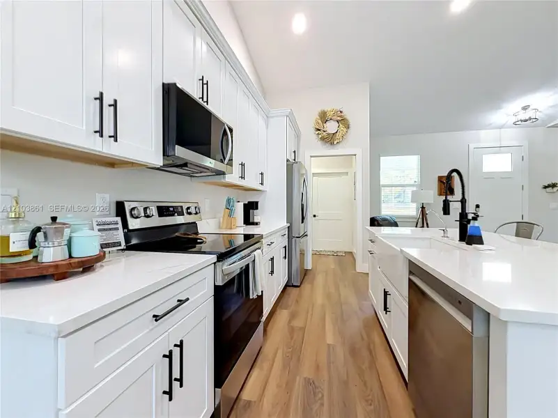 Kitchen featuring white shaker-style cabinetry, quartz countertops, stainless steel appliances, a farmhouse-style apron sink with a matte black faucet, and wood-finish flooring