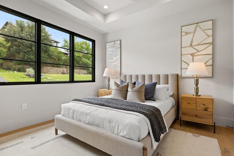 Bright bedroom featuring a tray ceiling with recessed lighting, black-framed windows, wood-finish flooring, and light-toned walls