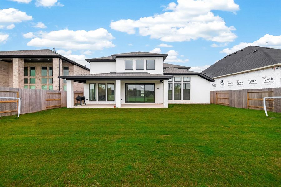This photo showcases the backyard of a modern, two-story home with a spacious lawn. The house features large windows, a covered patio, and is flanked by wooden fencing. Ideal for outdoor activities and entertaining.