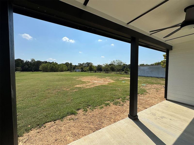 Covered patio with concrete flooring, black support columns, and a ceiling fan, overlooking an expansive grassy area with mature trees in the distance