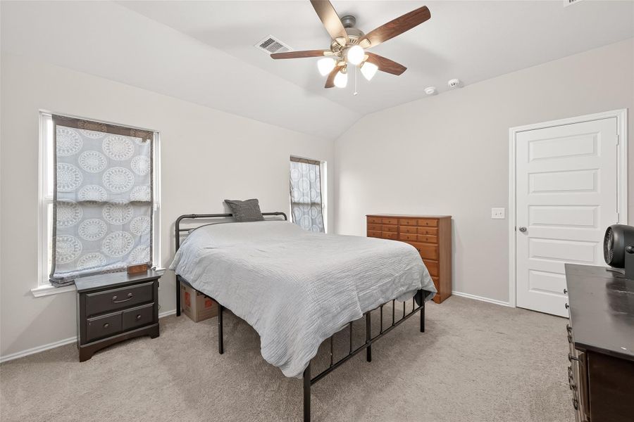 This is a cozy bedroom with neutral walls, carpeted floor, and a ceiling fan. It features a bed with metal frame, two windows with patterned curtains, and ample storage with a dresser and nightstand.