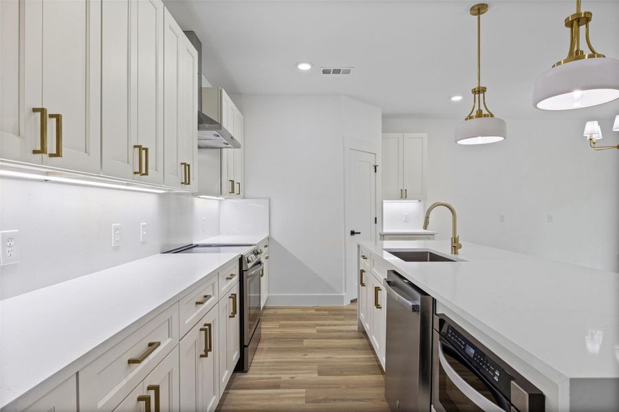 Modern kitchen featuring quartz countertops and backsplash, white shaker cabinetry with brass hardware, undermount sink with a brass pull-down faucet, stainless steel appliances, and light wood-finish flooring Modern kitchen featuring quartz countertops and backsplash, white shaker cabinetry with brass hardware, undermount sink with a brass pull-down faucet, stainless steel appliances, and light wood-finish flooring
