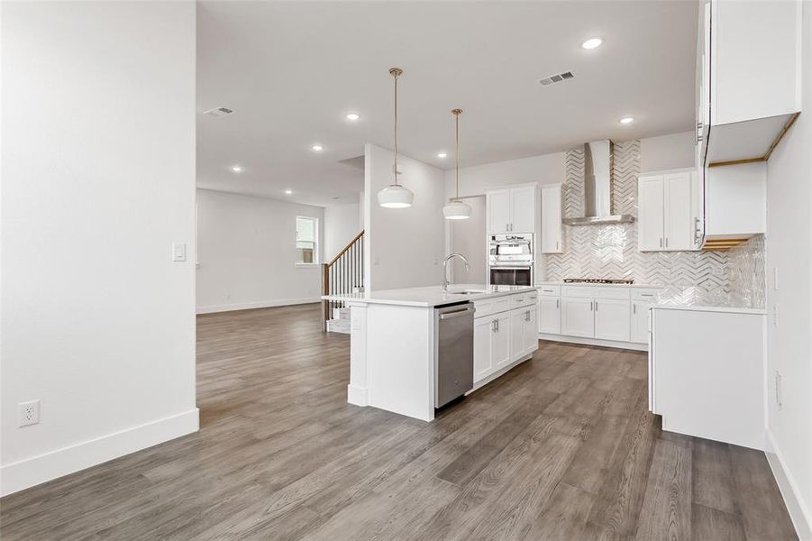 Kitchen featuring backsplash, a kitchen island with sink, wall chimney range hood, pendant lighting, and white cabinetry