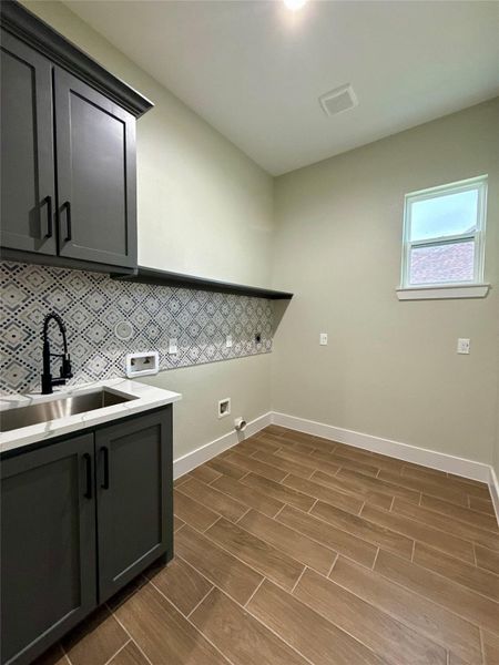 This photo shows a modern utility room with dark cabinetry, a stainless-steel sink, and patterned backsplash. The room features wood-look tile flooring and a small window, providing natural light. Ideal for laundry or additional storage.