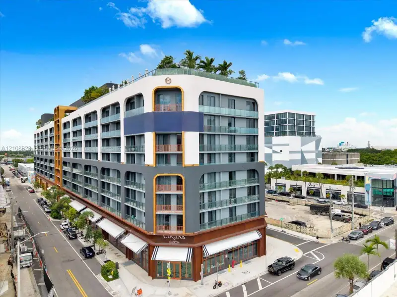 Modern multi-story structure featuring a rooftop terrace, glass-railed balconies, ground-level retail space with fabric awnings, and a dual-tone masonry facade