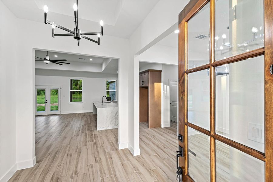This photo showcases a modern, open-concept living space with light wood-look tile flooring, a stylish chandelier, and large windows. The kitchen features a quartz waterfall island and contemporary fixtures. The area is bright and welcoming, with views of greenery through the glass doors. This photo showcases a modern, open-concept living space with light wood-look tile flooring, a stylish chandelier, and large windows. The kitchen features a quartz waterfall island and contemporary fixtures. The area is bright and welcoming, with views of greenery through the glass doors.
