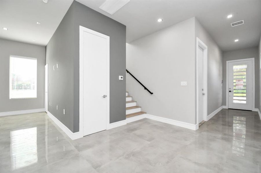 This is a modern, bright entryway featuring polished tile flooring, neutral gray and white walls, and ample natural light. There's a staircase with a sleek handrail leading to the upper level, two doors for additional rooms, and the main entrance door with glass panels.