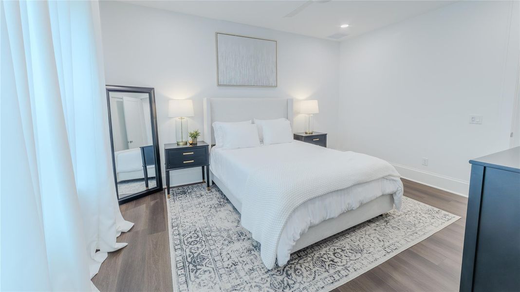 This bedroom features a bright, minimalist design with a cozy bed, dark wood flooring, and a stylish area rug. It includes two bedside tables with lamps, a full-length mirror, and a piece of wall art, creating a serene and inviting atmosphere.