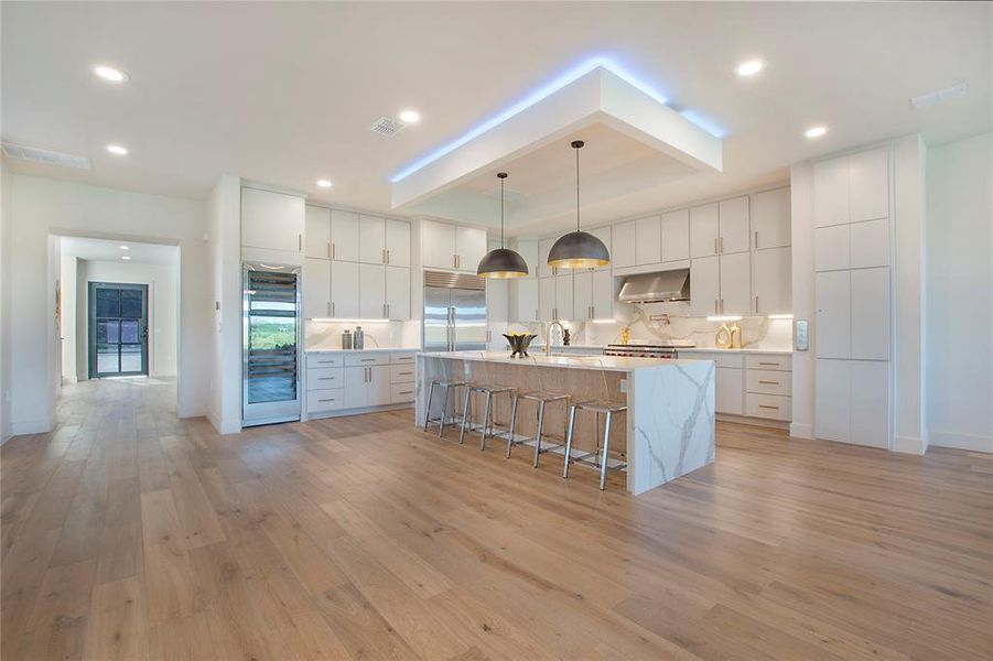 Kitchen with light hardwood / wood-style floors, hanging light fixtures, stainless steel built in fridge, and an island with sink