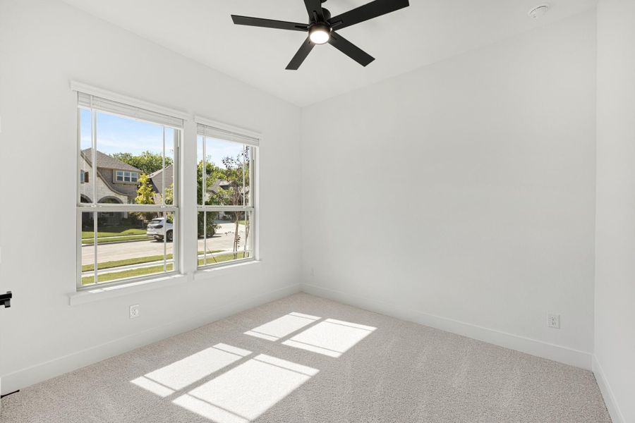 Empty bedroom with carpet flooring, black ceiling fan, and two windows overlooking a neighborhood street
