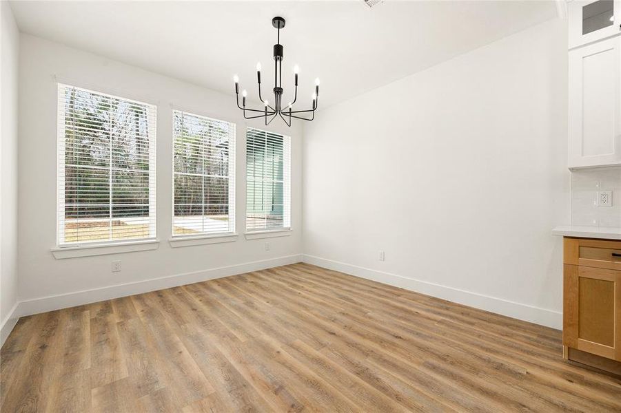 Bright dining area with large windows, modern chandelier, and wood-style flooring. Neutral walls and adjacent to a kitchen space.