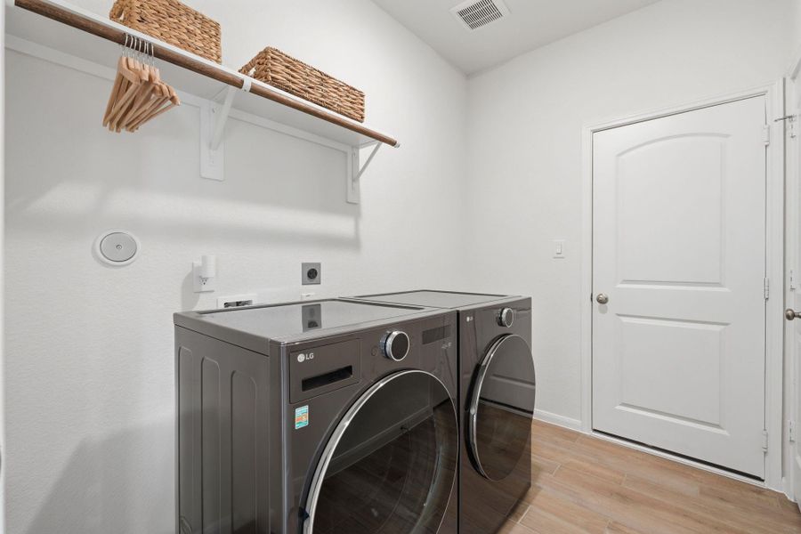 This laundry room features modern appliances, bright white walls, and light wood tile flooring. It includes a shelf with baskets and hangers for added storage and organization.