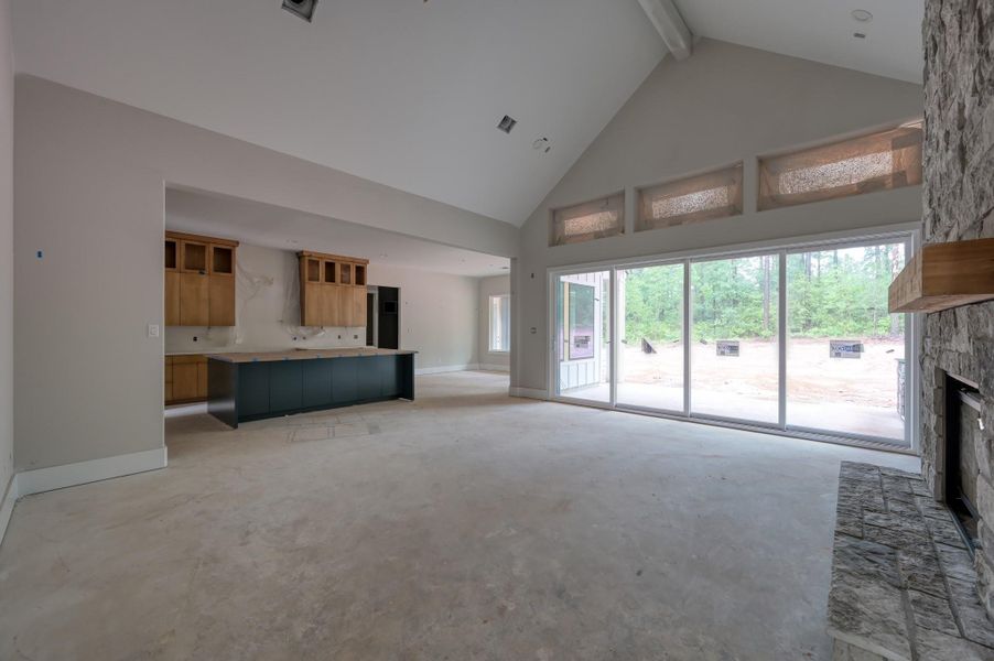 This spacious, open-concept living area features large windows for natural light, a stone fireplace, and a modern kitchen with an island. The vaulted ceiling adds to the airy feel, and sliding glass doors lead to a view of the wooded backyard and outdoor kitchen.