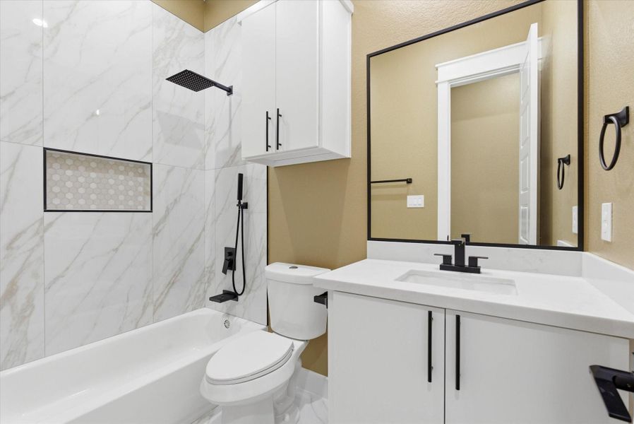 Hallway bathroom features a shower/tub combo with tile extending along the shower walls and matching tile flooring for an elevated look. It features both a handheld and standard showerhead, along with custom cabinetry for added storage and modern black finishes throughout.