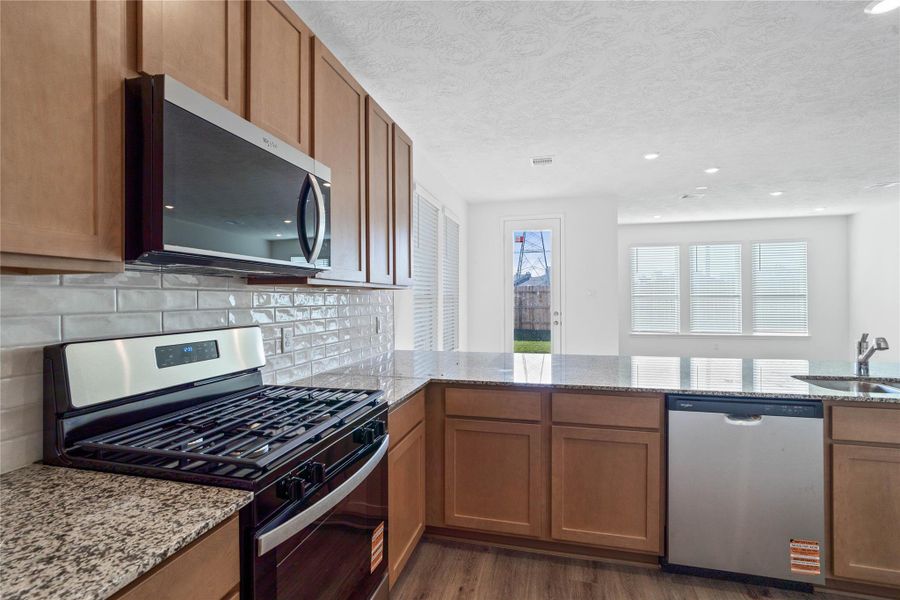This spacious kitchen boasts soaring ceilings, Woodmont Dakota cabinets in a Barley stain, granite countertops, stainless steel Whirlpool appliances, a modern Daltile backsplash, recessed lighting, an extended breakfast bar countertop, and a pantry—all overlooking the expansive family room.