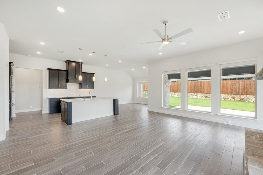 Open-concept living area with kitchen island, dark cabinets, wood-look tile floors, and large windows overlooking backyard. Open-concept living area with kitchen island, dark cabinets, wood-look tile floors, and large windows overlooking backyard.