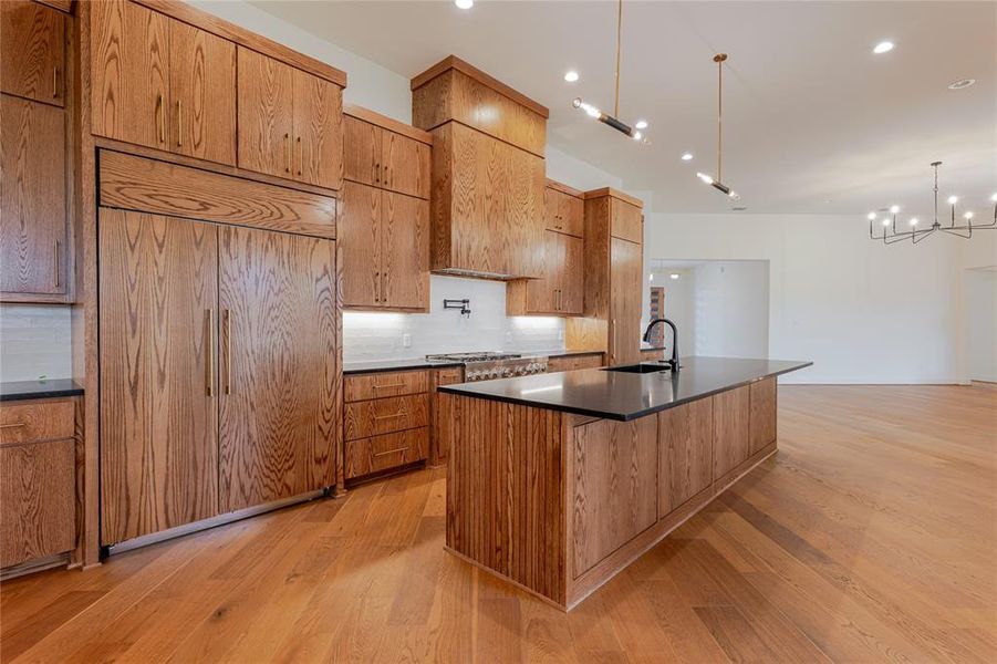 Kitchen featuring paneled built in refrigerator, an island with sink, pendant lighting, light wood finished floors, and decorative backsplash