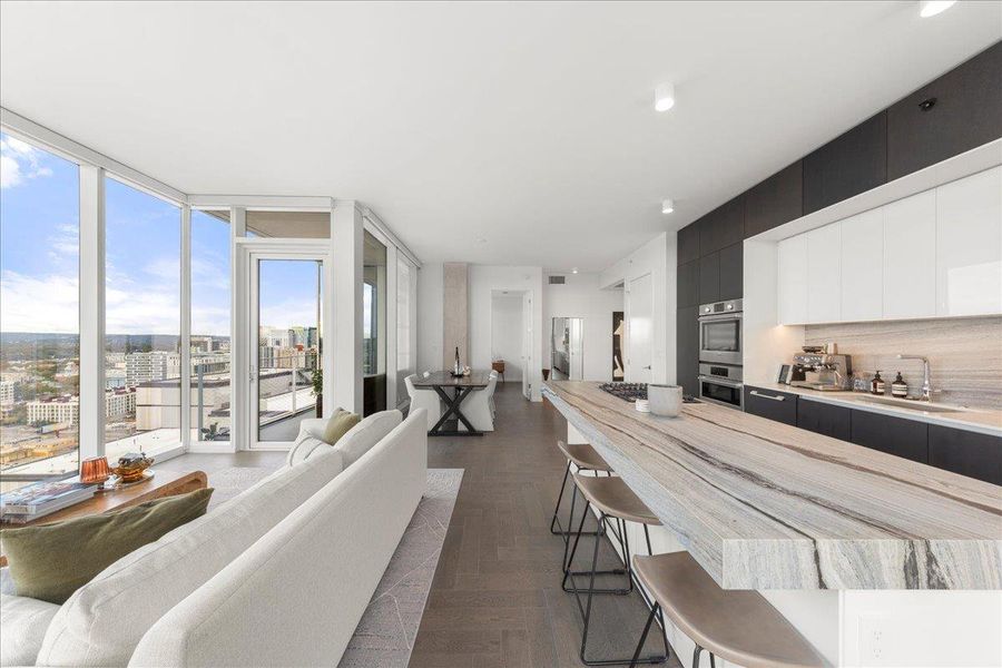 A designer kitchen anchored by upgraded Fantasy Ocean marble countertops and custom cabinetry, connecting directly to a light-filled living space with sweeping skyline vistas.