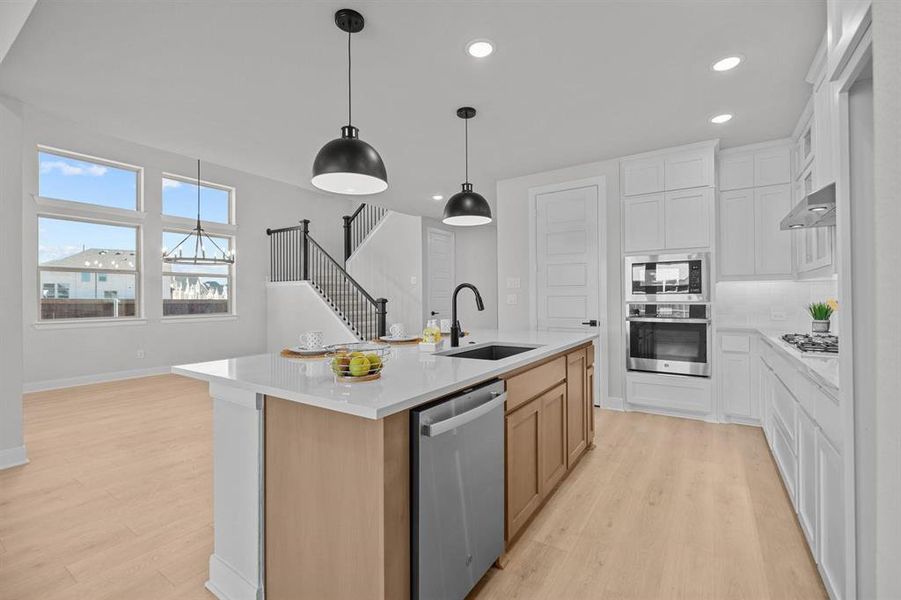 Open-concept kitchen featuring a central island with a built-in sink, stainless steel appliances, white cabinetry, and wood-finish flooring Open-concept kitchen featuring a central island with a built-in sink, stainless steel appliances, white cabinetry, and wood-finish flooring