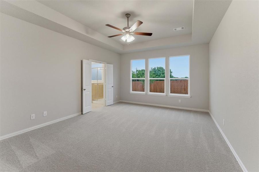 Spacious room featuring light-toned carpeting, a tray ceiling with a built-in ceiling fan, and three large windows providing abundant natural light