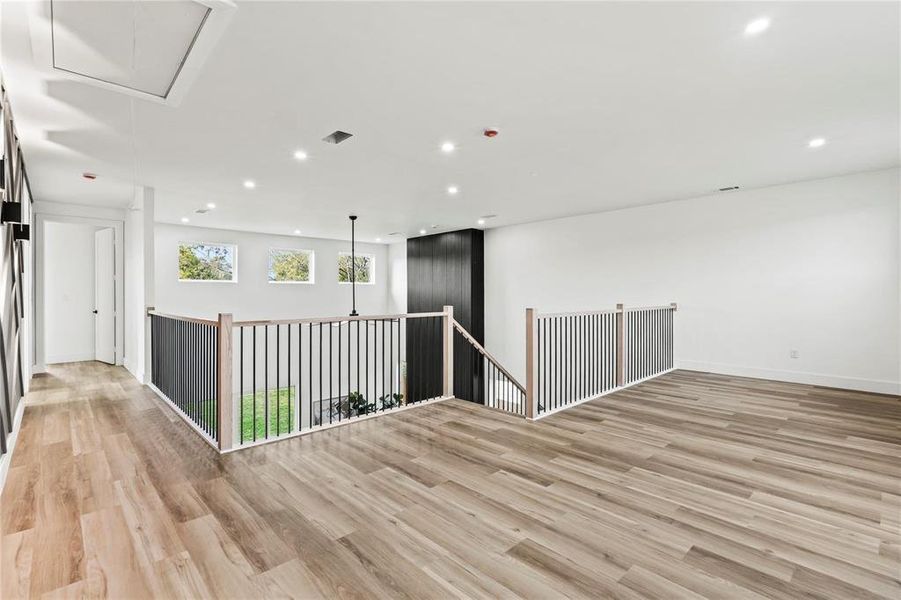 Spacious open loft area featuring wood-finish flooring, recessed lighting, and a contemporary railing with black spindles and a light wood-tone handrail