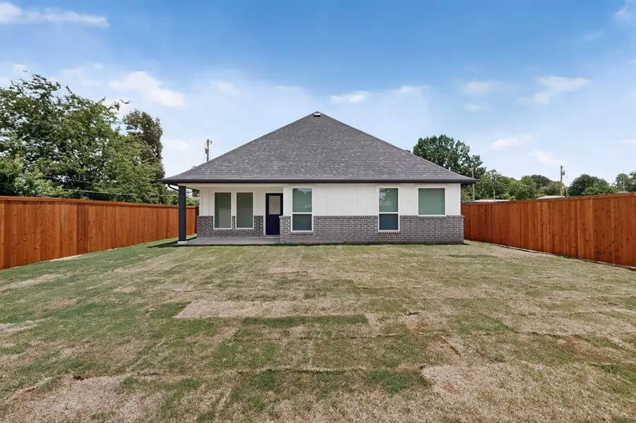 Expansive rear lawn featuring a privacy fence, covered patio, gray brick wainscoting, white siding, and a dark shingle roof