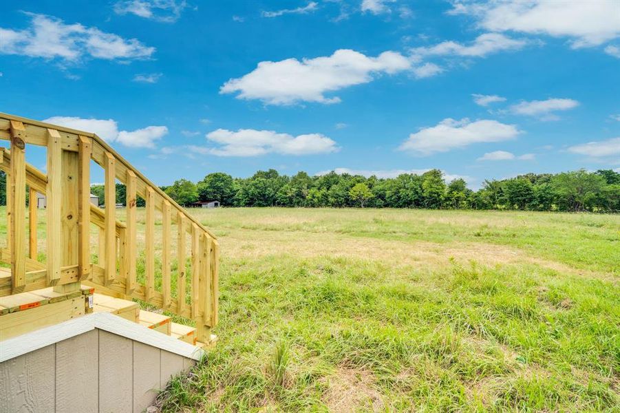 New construction wooden deck with balustrade and steps overlooking an expansive grassy field bordered by mature trees