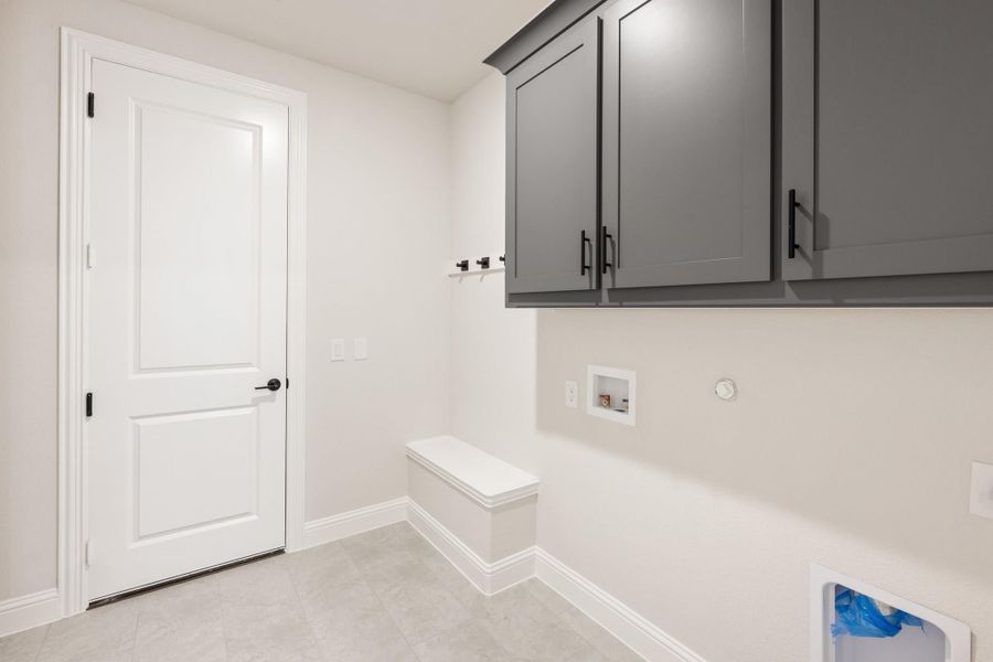 Laundry room with gray upper cabinets, built-in bench, wall hooks, and washer hookup on light tile floor