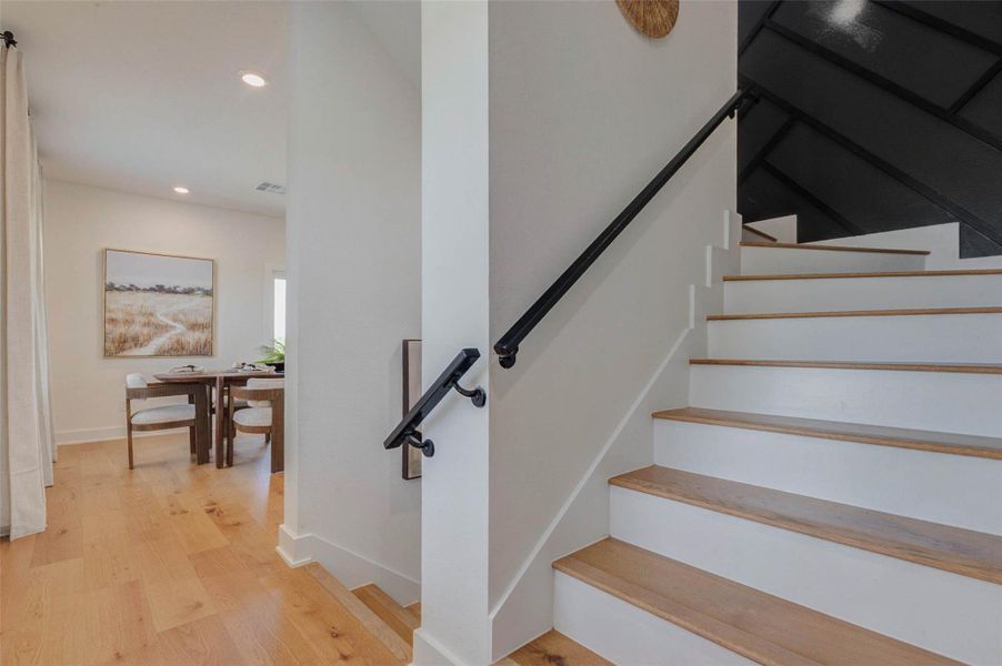 The staircase showcases a clean and modern design with wood treads, white risers, and sleek black handrails that create a striking contrast. Positioned conveniently within the home, it enhances both functionality and visual appeal. The open feel and quality finishes contribute to the home’s contemporary style and seamless flow between levels. The picture shown is from the model home with the same floor plan, Rev floor plan.