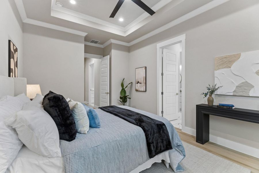Elegant tray ceilings and recessed lighting illuminate this tranquil bedroom, featuring light wood flooring and crisp white trim.