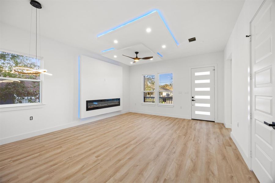 This modern living room features sleek wood-like flooring and a built-in electric fireplace. The ceiling has recessed lighting with blue accents and a stylish fan. Large windows and a contemporary front door brighten the space with natural light.
