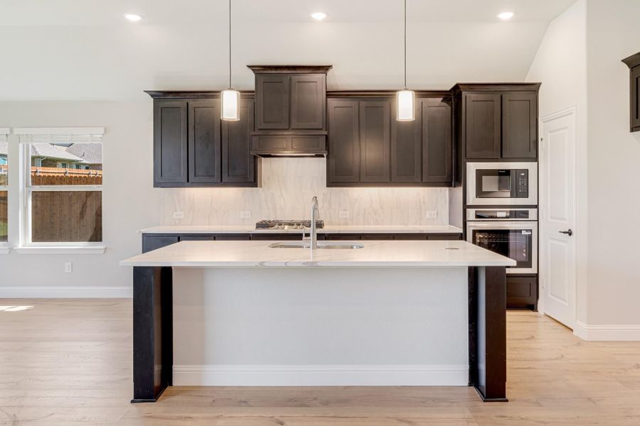 Kitchen with dark cabinets, white quartz island, marble backsplash, and built-in stainless steel appliances on light hardwood floors Kitchen with dark cabinets, white quartz island, marble backsplash, and built-in stainless steel appliances on light hardwood floors