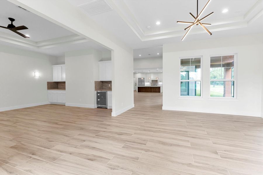 This spacious room features sleek light wood flooring and a modern open layout. It includes built-in white cabinetry with a stone backsplash, recessed lighting, and a ceiling fan. Large windows offer plenty of natural light and outdoor views.