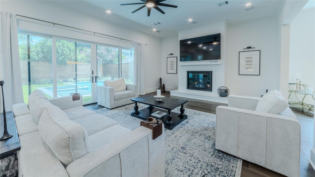 This spacious living room features a modern design with a cozy fireplace, large wall-mounted TV, and elegant furnishings. Sliding glass doors provide ample natural light and lead to a serene backyard with a pool, creating a seamless indoor-outdoor living experience.