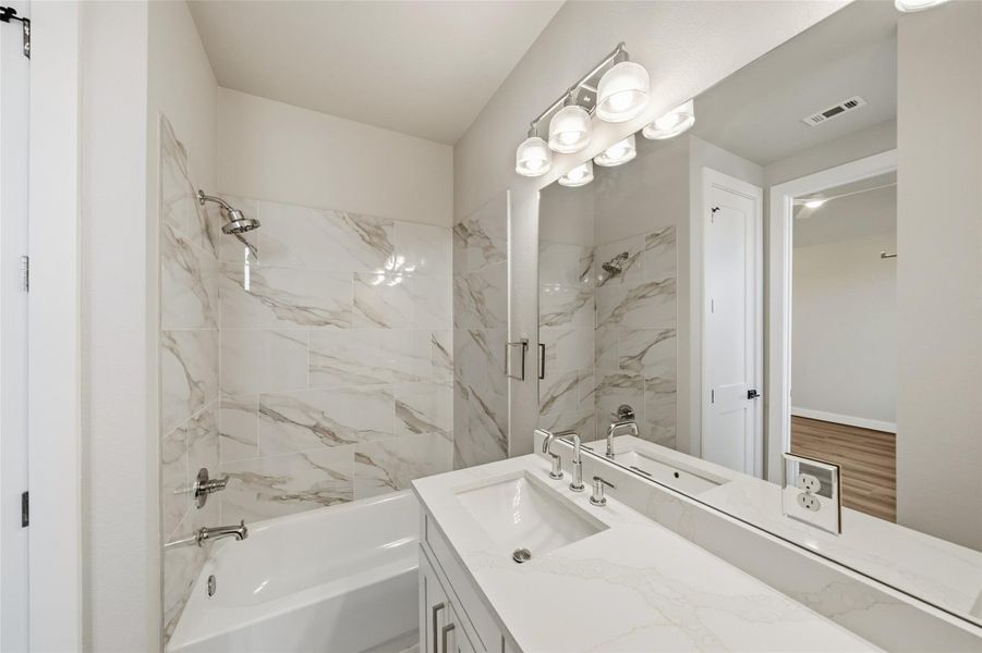 This bathroom features a modern design with a marble-patterned tile surround for the bathtub and shower combo, a sleek vanity with a white countertop and dual sinks, and a large mirror under stylish lighting.
