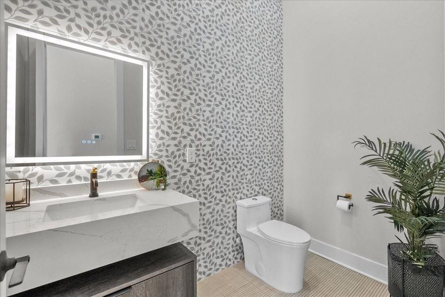 This bathroom features a stylish leaf-patterned accent wall, a modern vanity with a sleek illuminated mirror, and elegant fixtures. A potted plant adds a touch of greenery. This bathroom features a stylish leaf-patterned accent wall, a modern vanity with a sleek illuminated mirror, and elegant fixtures. A potted plant adds a touch of greenery.