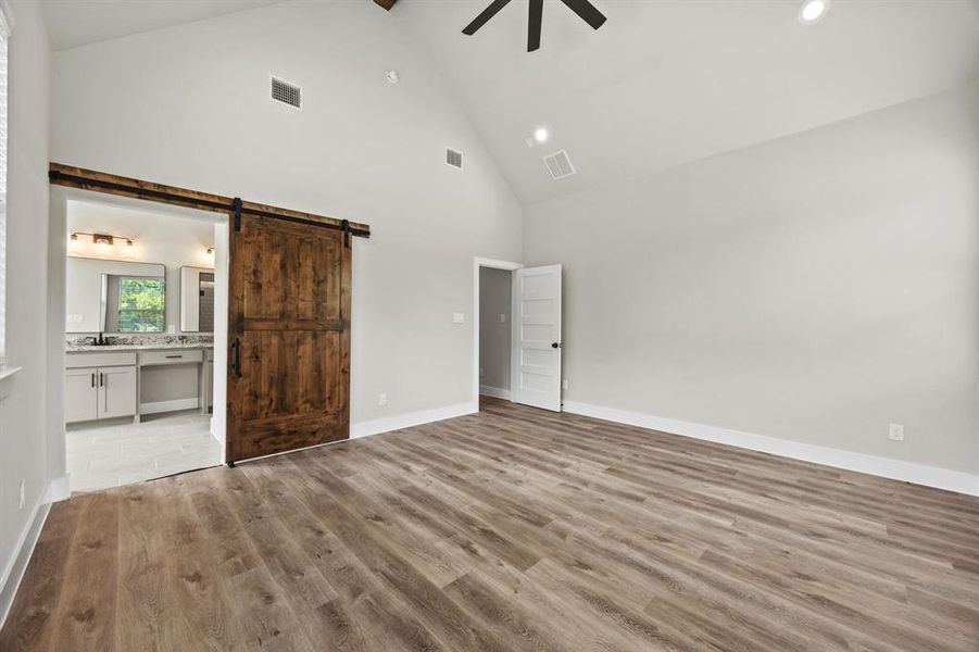 Unfurnished bedroom with high vaulted ceiling, a barn door, light wood finished floors, ceiling fan, and ensuite bath Unfurnished bedroom with high vaulted ceiling, a barn door, light wood finished floors, ceiling fan, and ensuite bath