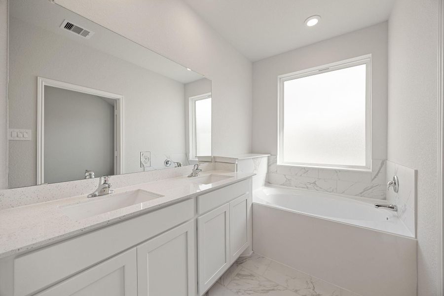 An immaculate and bright primary bathroom retreat featuring a dual vanity with crisp white cabinetry and quartz countertops, under a large frameless mirror. The space includes a spacious built-in soaking tub with a sleek white marble-look tile surround and a privacy window, all complemented by elegant marble-look tile flooring and modern fixtures, creating a spa-like atmosphere. An immaculate and bright primary bathroom retreat featuring a dual vanity with crisp white cabinetry and quartz countertops, under a large frameless mirror. The space includes a spacious built-in soaking tub with a sleek white marble-look tile surround and a privacy window, all complemented by elegant marble-look tile flooring and modern fixtures, creating a spa-like atmosphere.