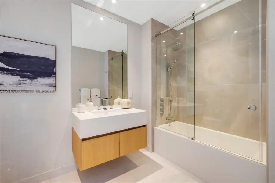 Floating wood-finish vanity with a rectangular sink, large format tile shower surround, glass sliding shower door, and wall-mounted oversized mirror Floating wood-finish vanity with a rectangular sink, large format tile shower surround, glass sliding shower door, and wall-mounted oversized mirror
